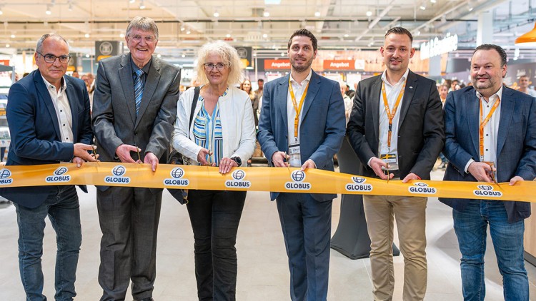 Sechs Personen halten ein gelbes Band mit GLOBUS-Logo in einem hell beleuchteten Einkaufsmarkt.