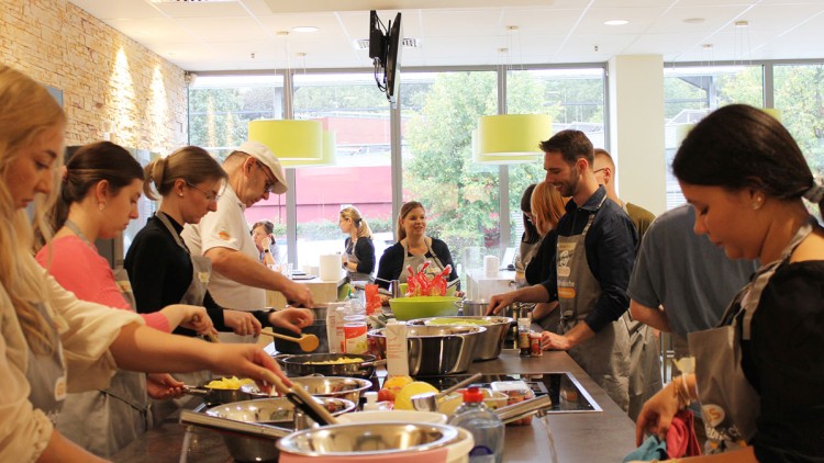 Mehrere Personen arbeiten an einer Küchenzeile mit Schüsseln, Pfannen und Zutaten, im Hintergrund sind Fenster und Lampen.