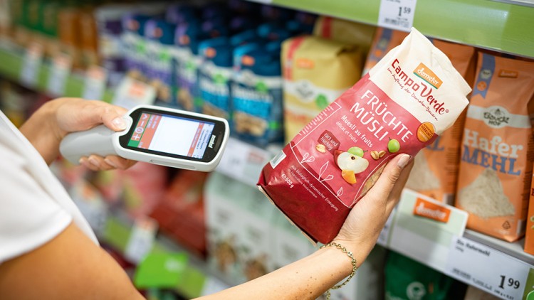 Person scannt mit Scan & Go-Gerät eine Packung rotes Früchte-Müsli der Marke Campo Verde im GLOBUS-Regal.
