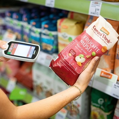 Person scannt mit Scan & Go-Gerät eine Packung rotes Früchte-Müsli der Marke Campo Verde im GLOBUS-Regal.