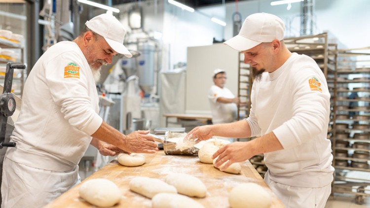 Zwei Personen in weißer Arbeitskleidung formen Teig auf einem Holztisch in einer hellen Bäckereiumgebung, GLOBUS-Logo sichtbar.