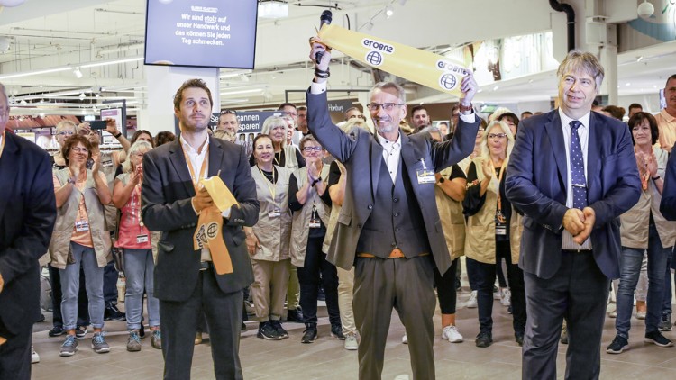 Person in Anzug hält ein gelbes Eröffnungsband mit GLOBUS-Logo hoch, umgeben von applaudierenden Menschen in einem Markt.