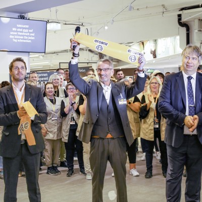 Person in Anzug hält ein gelbes Eröffnungsband mit GLOBUS-Logo hoch, umgeben von applaudierenden Menschen in einem Markt.