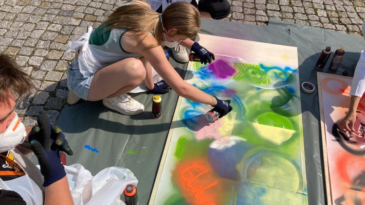 Personen gestalten mit bunten Sprühfarben Schablonen und kreative Motive auf großes Papier, „Globus“ steht in Orange darauf.