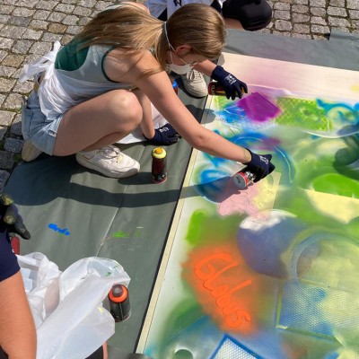 Personen gestalten mit bunten Sprühfarben Schablonen und kreative Motive auf großes Papier, „Globus“ steht in Orange darauf.