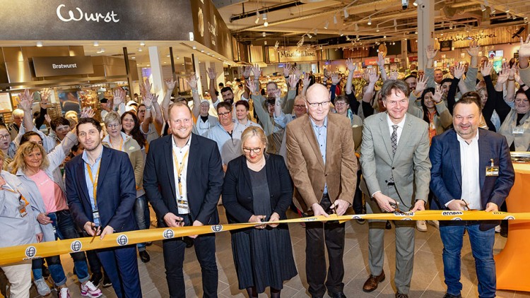 Eine Gruppe von Menschen steht vor einer Absperrung mit GLOBUS-Logo, im Hintergrund eine Wursttheke und ein Geschäft.