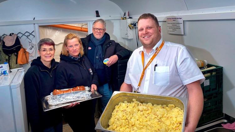 Ein Mensch in weißem Hemd hält ein Tablett mit Kartoffelsalat, daneben wird ein Tablett mit Wurst präsentiert.