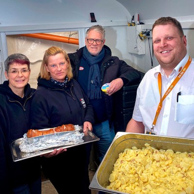 Ein Mensch in weißem Hemd hält ein Tablett mit Kartoffelsalat, daneben wird ein Tablett mit Wurst präsentiert.