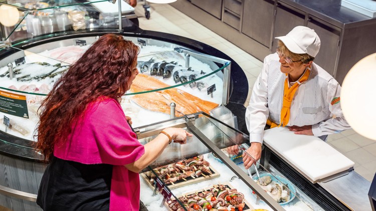 Kundin zeigt auf frischen Fisch und Meeresfrüchte in einer Theke, während ein Mitarbeitender in weißer Arbeitskleidung berät.