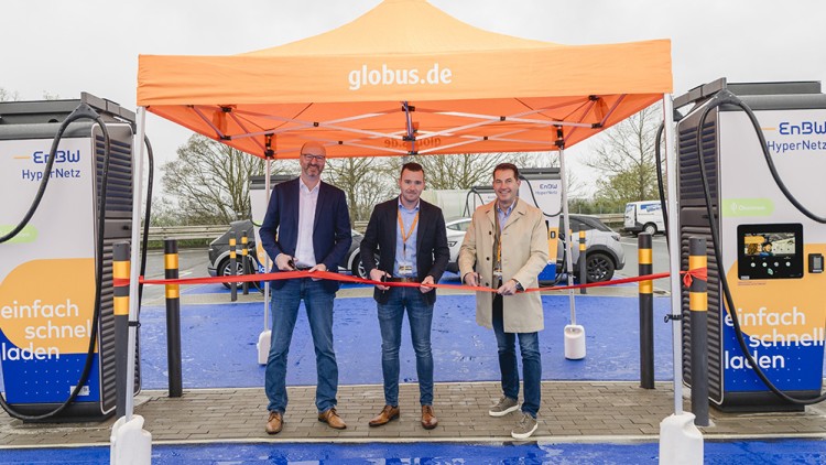 Drei Personen schneiden ein rotes Band unter einem orangefarbenen Pavillon mit der Aufschrift globus.de durch.