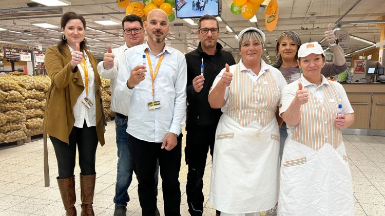 Eine Gruppe von Personen steht in einem GLOBUS-Markt vor Kartoffelstapeln, zeigt Daumen hoch und hält blaue Kugelschreiber.