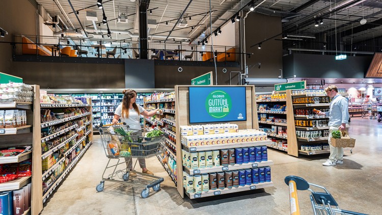 Innenansicht eines GLOBUS-Marktes, Frau mit Einkaufswagen vor Regalen, Display mit „GUTfühl MARKT“ in der Mitte.