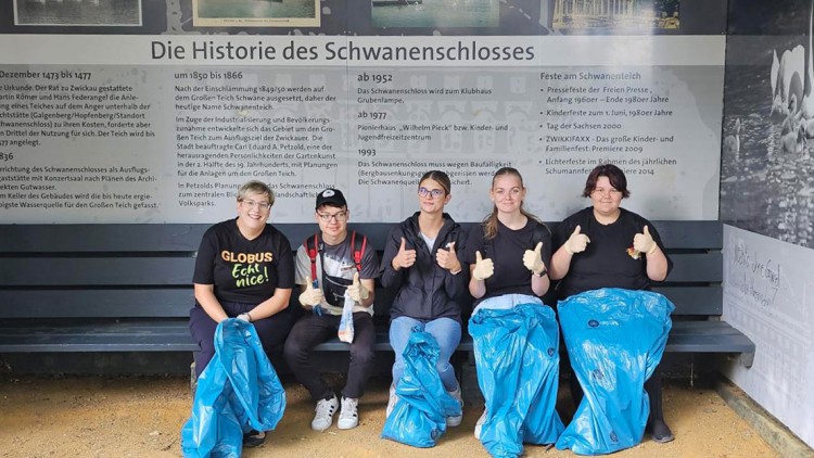 Fünf Personen sitzen auf einer Bank vor einer Infotafel zur Geschichte des Schwanenschlosses mit blauen Müllsäcken und Handschuhen.