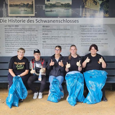 Fünf Personen sitzen auf einer Bank vor einer Infotafel zur Geschichte des Schwanenschlosses mit blauen Müllsäcken und Handschuhen.