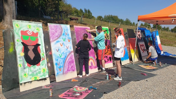 Mehrere Personen gestalten mit Farbspraydosen bunte Motive auf großen aufgestellten Holzbrettern im Freien.
