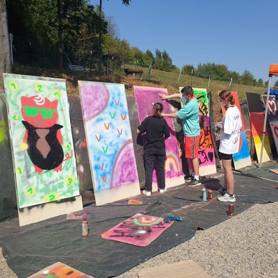 Mehrere Personen gestalten mit Farbspraydosen bunte Motive auf großen aufgestellten Holzbrettern im Freien.
