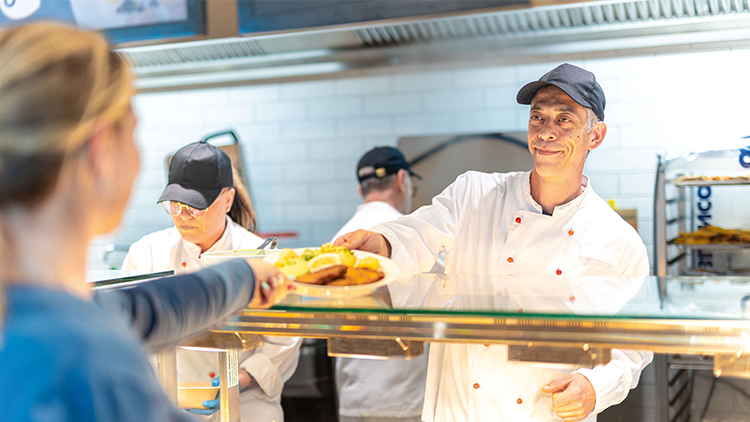 Eine Person in weißer Kochkleidung reicht hinter einer Theke einen Teller mit frittierten Speisen und Beilage an eine andere Person.