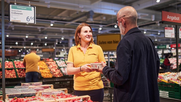 Eine Person im gelben Polohemd überreicht einem Kunden eine Erdbeerschale, im Hintergrund Obstkisten und Angebotschilder.