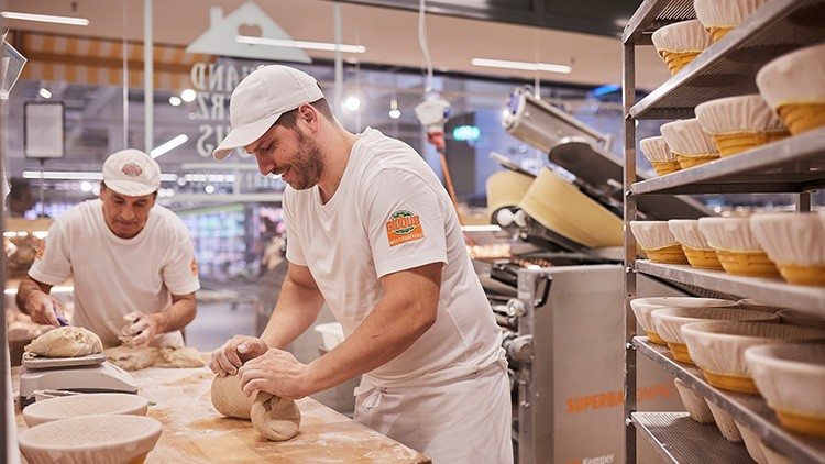 Zwei Bäcker in weißer Kleidung formen Brotteig, im Hintergrund sind Backutensilien und Regale mit Gärkörben zu sehen.