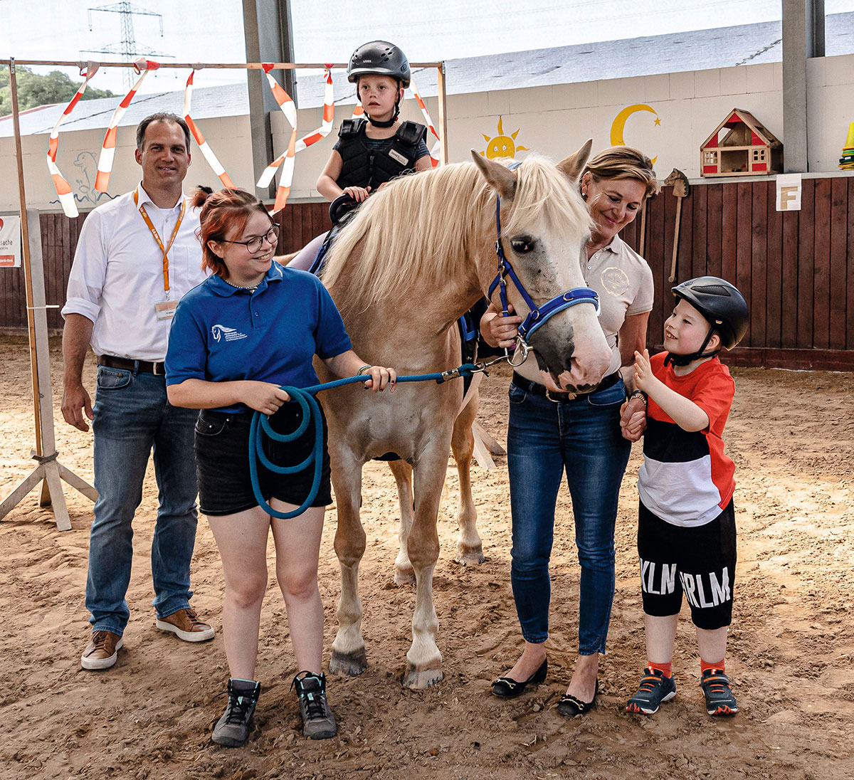 Ein Pferd mit einem blauen Halfter steht in einer Gruppe von Menschen auf einem Sandplatz in einer Halle.