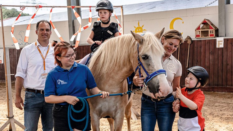 Ein Pferd mit einem blauen Halfter steht in einer Gruppe von Menschen auf einem Sandplatz in einer Halle.