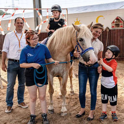 Ein Pferd mit einem blauen Halfter steht in einer Gruppe von Menschen auf einem Sandplatz in einer Halle.