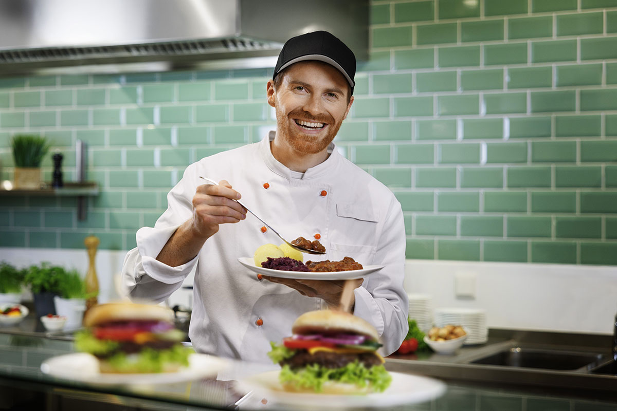 Koch in weißer Uniform hält einen Teller mit Fleisch, Rotkohl und Kartoffeln, grüne Fliesenwand und Zutaten im Hintergrund.