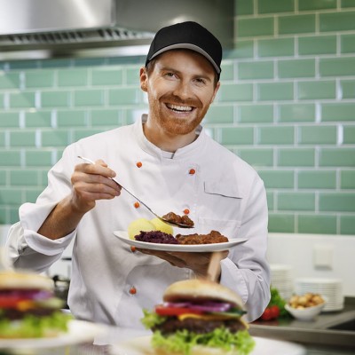 Koch in weißer Uniform hält einen Teller mit Fleisch, Rotkohl und Kartoffeln, grüne Fliesenwand und Zutaten im Hintergrund.