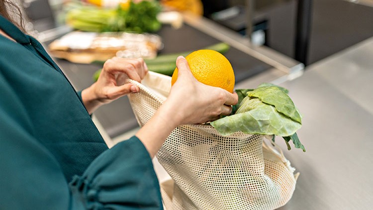 Person legt eine Orange und Kohlblätter in eine weiße Stofftasche, daneben liegen Gemüse und Brot auf einer Verkaufstheke.