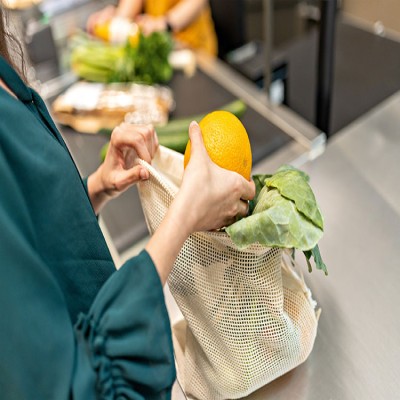 Person legt eine Orange und Kohlblätter in eine weiße Stofftasche, daneben liegen Gemüse und Brot auf einer Verkaufstheke.