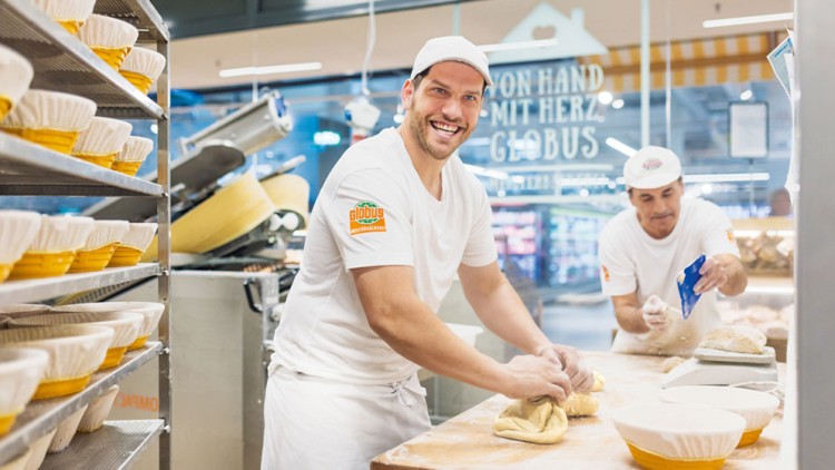 Bäcker formen Teig auf einer Arbeitsfläche, gelbe Schalen stehen daneben, im Hintergrund Schriftzug „Von Hand mit Herz bei GLOBUS“.