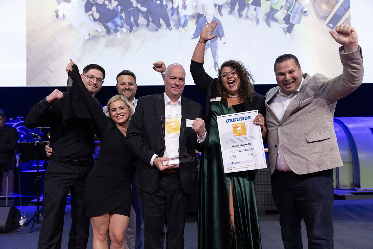 Fünf Menschen stehen mit einer Urkunde und einem Glasaward vor einer Leinwand, feiern und heben die Arme.