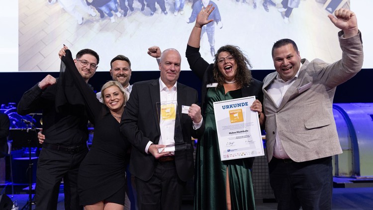 Fünf Menschen stehen mit einer Urkunde und einem Glasaward vor einer Leinwand, feiern und heben die Arme.