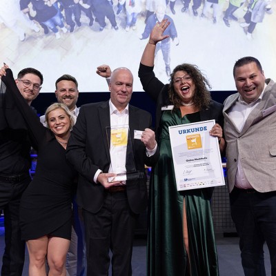 Fünf Menschen stehen mit einer Urkunde und einem Glasaward vor einer Leinwand, feiern und heben die Arme.