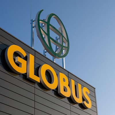 Gelbes GLOBUS-Logo auf einer dunklen Gebäudefassade, darüber grüne Globus-Skulptur vor blauem Himmel.