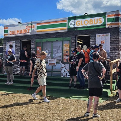 Menschen stehen vor einem GLOBUS Baumarkt-Stand mit Werbebannern, grünen Stufen und Informationsschildern.