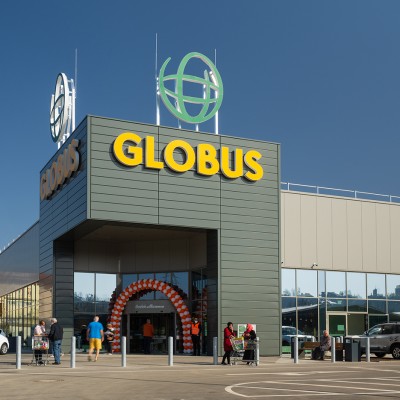 Eingangsbereich eines GLOBUS Marktes mit gelbem Schriftzug, orangenen und weißen Luftballons sowie Menschen mit Einkaufswagen.