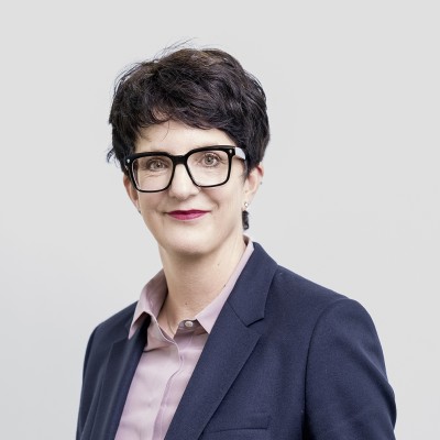 Person mit kurz geschnittenem dunklem Haar trägt ein dunkelblaues Sakko und ein hellrosa Hemd vor grauem Hintergrund.