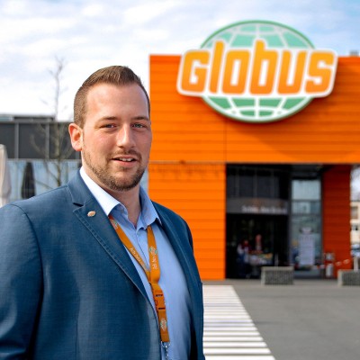 Eine Person in einem blauen Anzug steht vor einem GLOBUS-Markt mit orangefarbener Fassade und dem Logo oben.
