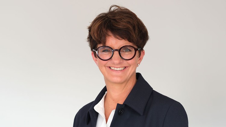 Person in dunkelblauem Mantel und weißem Hemd vor hellem Hintergrund, Gesicht unkenntlich gemacht.