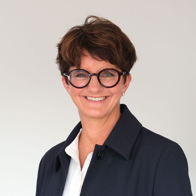 Person in dunkelblauem Mantel und weißem Hemd vor hellem Hintergrund, Gesicht unkenntlich gemacht.