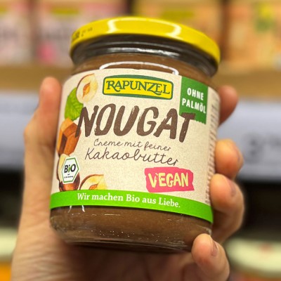 Ein Glas „Rapunzel Nougat Creme mit feiner Kakaobutter“ wird von einer Hand gehalten, mit den Labels Bio, vegan und ohne Palmöl.
