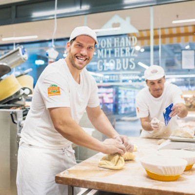Bäcker in weißer Kleidung formen Teig auf einer bemehlten Arbeitsfläche, daneben Körbe und ein Schriftzug „Von Hand mit Herz bei GLOBUS“.