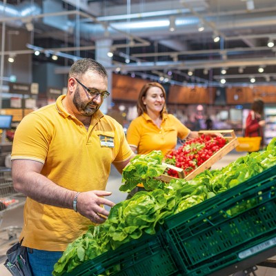 Mitarbeitende in gelber Arbeitskleidung sortieren rote Radieschen und grünen Salat in einem Markt vor Regalen und Einkaufswagen.