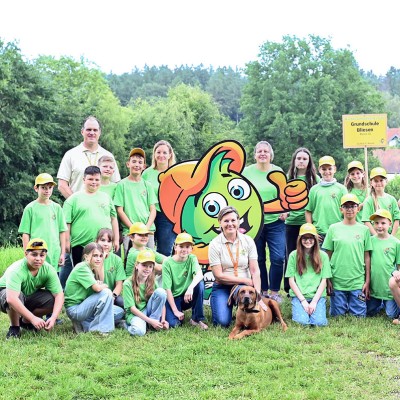 Menschen und Globini stehen und sitzen auf einer Wiese vor einem Schild mit der Aufschrift „Grundschule Bliesen“.