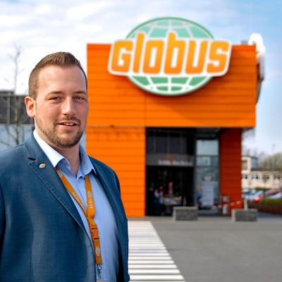 Person in blauem Anzug mit orangefarbenem GLOBUS-Schlüsselband vor einem GLOBUS-Markt mit orangener Fassade.