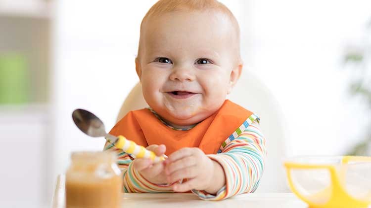 Kind in gestreiftem Langarmshirt und orangener Lätzchen hält einen gelben Löffel, vor ihm steht ein Glas mit Brei.