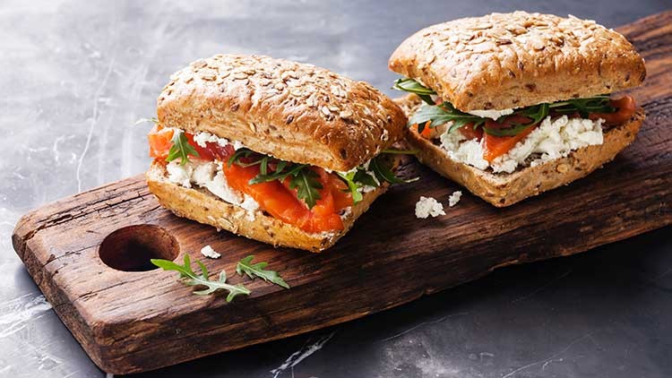 Zwei Körnerbrötchen mit Lachs, Frischkäse und Rucola auf einem hölzernen Schneidebrett.