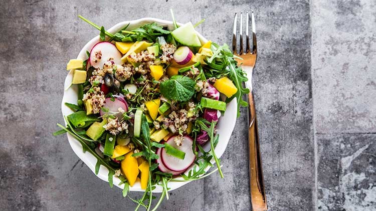 Bunte Schüssel mit Salat, Quinoa, Radieschen, Mango, Gurke und Rucola, daneben liegt eine silberne Gabel auf grauem Untergrund.