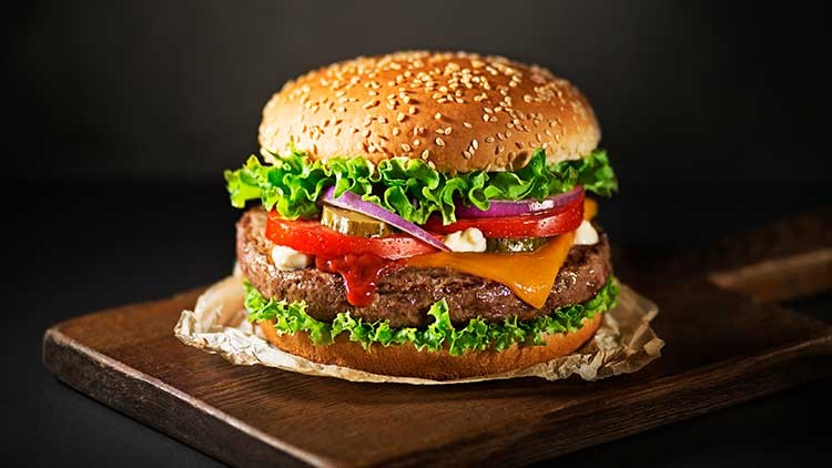 Burger mit Sesambrötchen, grünem Salat, Rindfleischpatty, Käsescheibe, Tomaten, Zwiebeln, Gurkenscheiben und Ketchup.
